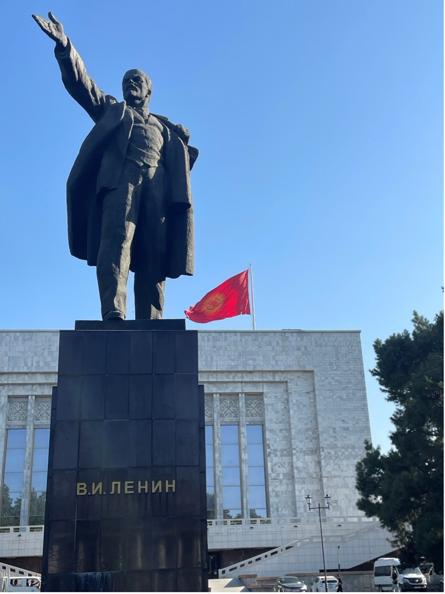 A monumental stone stature of Lenin standing upright with arm raised and outstretched, gesturing palm up towards the sky