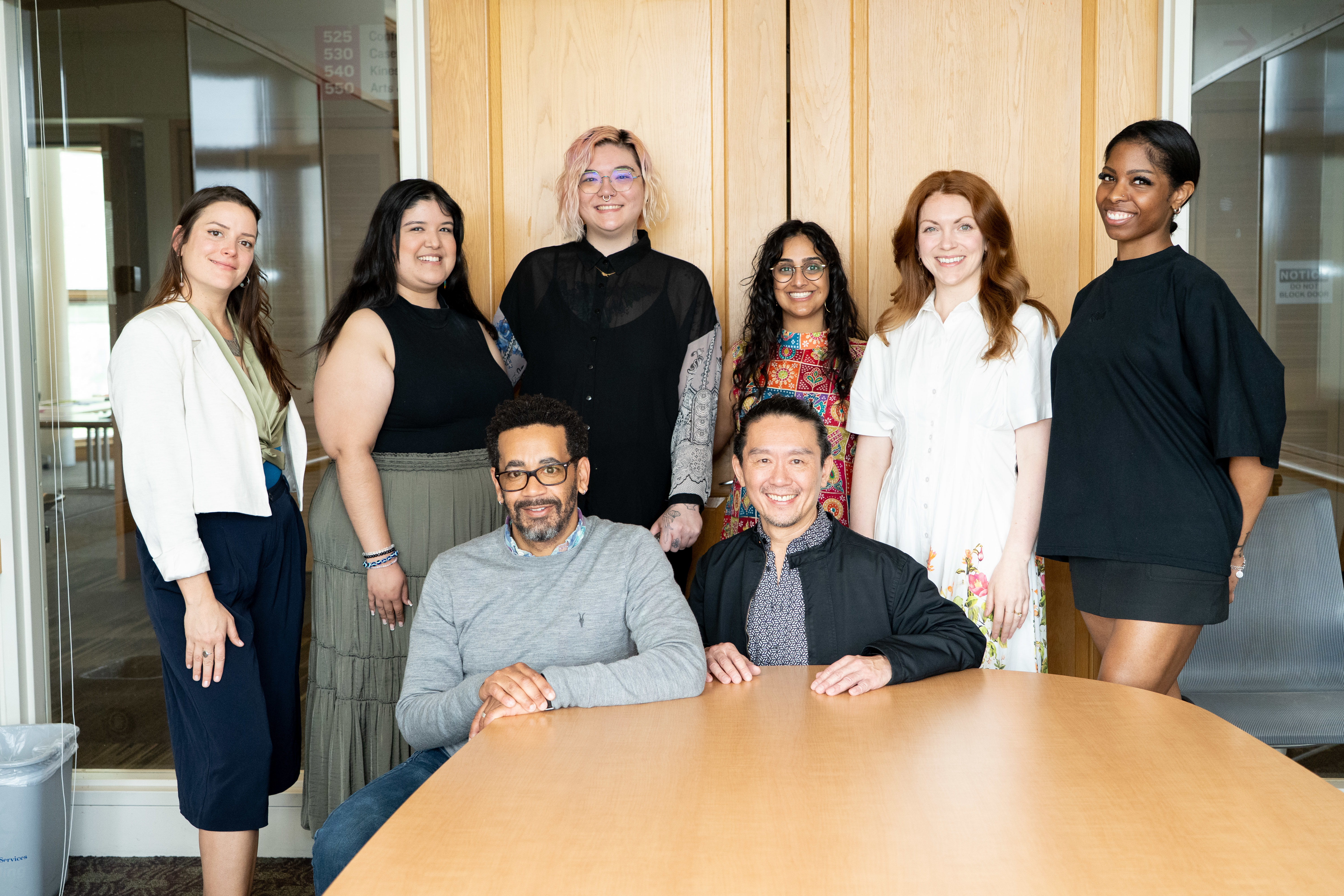 Students and faculty smile sitting and standing at board room table